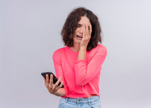 Foto gratuita destacó joven hermosa mujer sosteniendo el teléfono móvil y poniendo la mano en la cara en la pared blanca aislada con espacio de copia