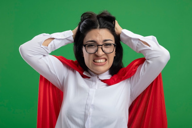 Destacó a joven superhéroe caucásica con gafas sosteniendo su cabeza y mostrando los dientes mirando a cámara aislada sobre fondo verde