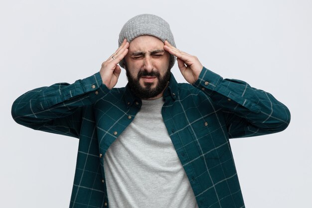 Destacó joven vistiendo sombrero de invierno manteniendo las manos en la cabeza con los ojos cerrados aislado sobre fondo blanco.