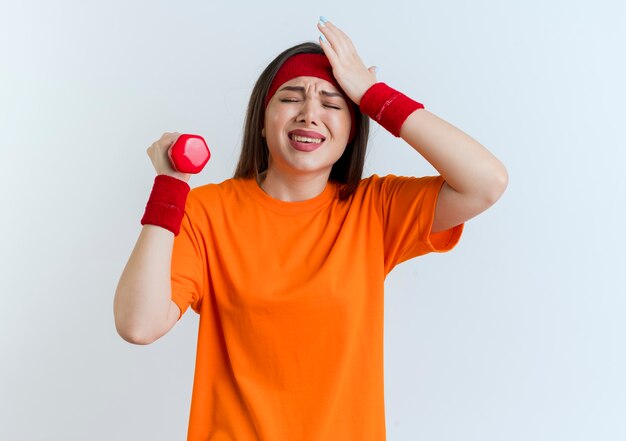 Destacó la mujer deportiva joven con diadema y muñequeras poniendo la mano en la cabeza sosteniendo mancuernas con los ojos cerrados aislados en la pared blanca