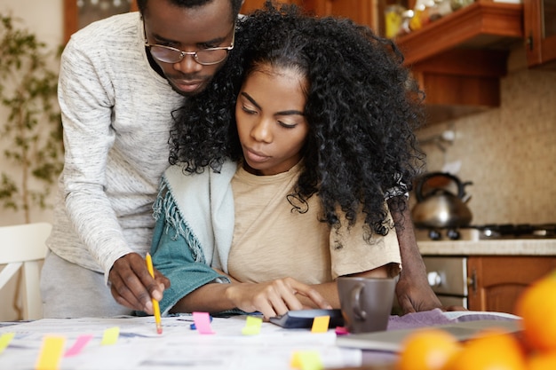 Destacó la pareja africana que tiene muchas deudas, haciendo trámites en casa. Hombre serio en gafas con lápiz apuntando a un trozo de papel sobre la mesa mientras su joven esposa hace cálculos en la calculadora