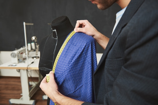 Detalle del talentoso diseñador profesional joven caucásico que trabaja en un vestido nuevo para la colección de primavera, revisa los tamaños con cinta métrica y pasa el día en su taller