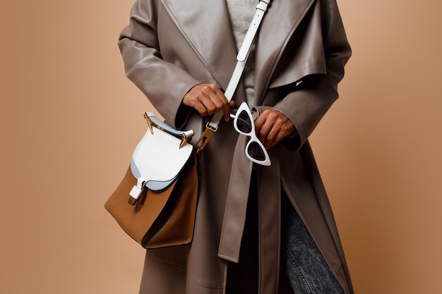Detalles Mujer negra con abrigo de cuero gris, posando sobre fondo beige. Bolso marrón y gafas de sol blancas en las manos. Concepto de moda de otoño o invierno.