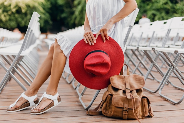 Detalles de primer plano de mujer con vestido blanco, sombrero rojo sentado en el teatro al aire libre de verano solo en una silla, tendencia de moda de estilo callejero de primavera, accesorios, viajar con mochila, piernas delgadas en sandalias