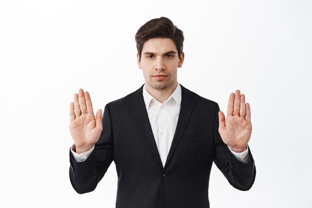 Detente, mantén la distancia. Hombre de negocios serio y decidido en traje negro muestra el bloque, rechaza o prohíbe el gesto, diga que no, prohíba algo malo, de pie sobre el fondo blanco