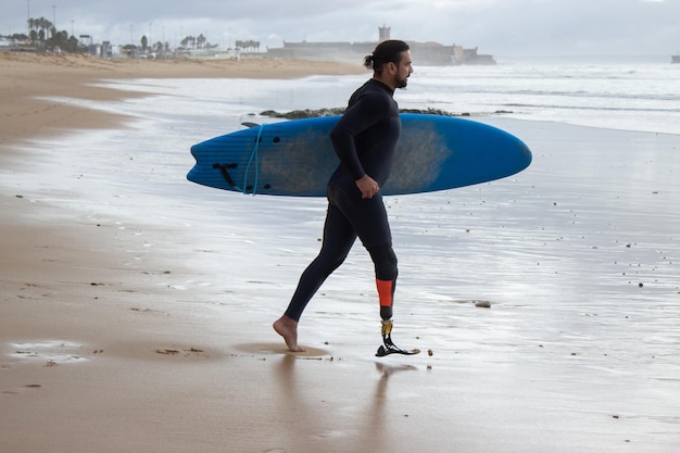 Determinado hombre de pelo oscuro entrenando solo en la playa. Surfista adulto medio con tabla de surf, caminando hacia el agua. Deporte, ocio, concepto de estilo de vida activo.