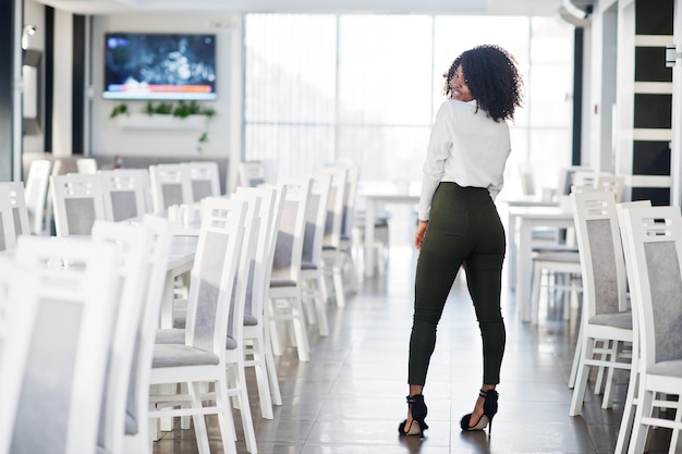 Detrás de la alegre y sexy dama afroamericana de negocios con cabello afro, usa blusa blanca y pantalones verdes posados en el café