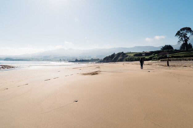 Detrás de la foto de una mujer caminando sobre la arena de la playa cerca de la orilla con montañas