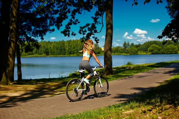 Detrás del modelo de mujer rubia sexy deporte caliente mujer montando en bicicleta en el parque verde de verano cerca del lago con el pelo elevado volando en el aire