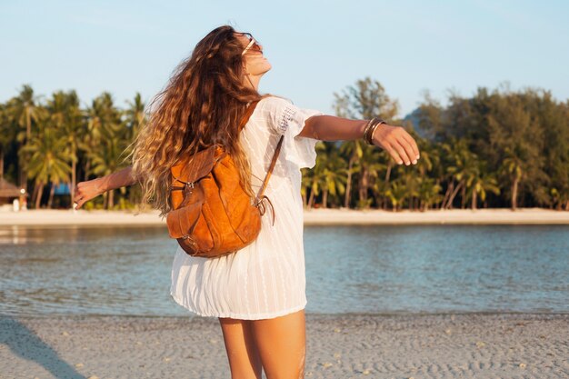 Detrás de una mujer hermosa en vestido blanco caminando sin preocupaciones en la playa tropical con mochila de cuero.