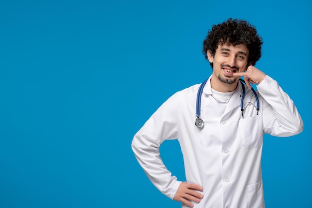 Día del doctor chico lindo guapo rizado en uniforme médico haciendo gesto de llamada telefónica