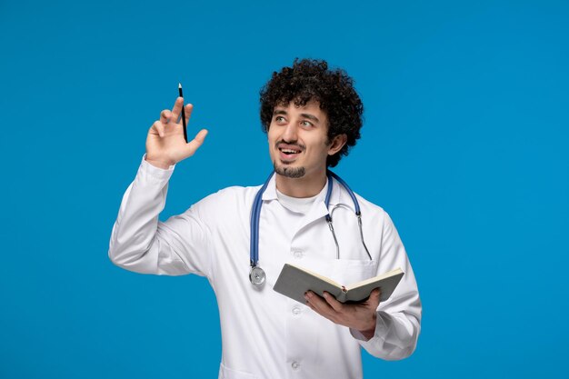 Día del doctor chico lindo guapo rizado en uniforme médico sonriendo y sosteniendo un bolígrafo con el cuaderno