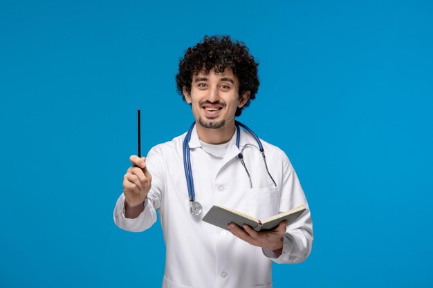 Día del doctor chico lindo guapo rizado en uniforme médico sonriendo y sosteniendo la pluma con el libro