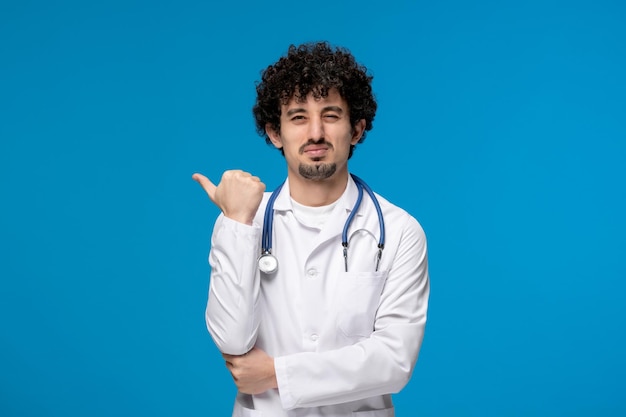 Día del doctor chico lindo moreno rizado en uniforme médico apuntando a la izquierda y haciendo cara de mal humor