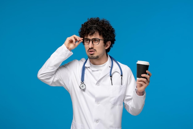 Día del doctor chico lindo moreno rizado en uniforme médico sosteniendo un vaso de papel y pensando