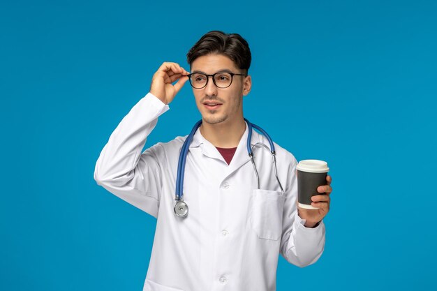 Día del doctor lindo joven moreno en bata de laboratorio con gafas y sosteniendo una taza de café