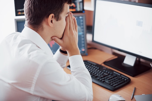 Foto gratuita día estresante en la oficina. hombre de negocios joven que lleva a cabo las manos en su cara mientras que se sienta en el escritorio en oficina creativa. concepto gráfico de la bolsa de comercio de forex finance