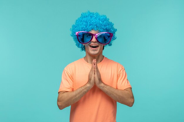 Día de los inocentes chico payaso emocionado manos juntas cabello azul rosa grandes gafas de sol divertido sonriendo feliz