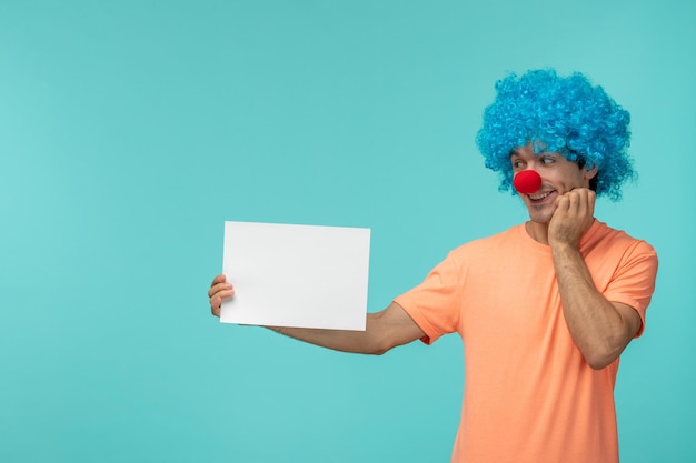 Día de los inocentes chico payaso feliz mano en la barbilla sosteniendo papel pelo azul emocionado gracioso nariz roja