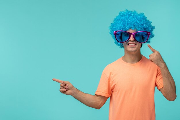 Día de los inocentes chico payaso sonriente cabello azul rosa grandes gafas de sol divertido feliz señalando las manos a la izquierda