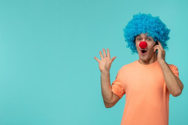 Día de los inocentes chico payaso sorprendido sosteniendo el teléfono cabello azul manos arriba emocionado gracioso nariz roja