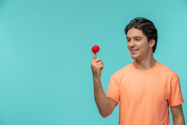 Día de los inocentes chico sonriendo sosteniendo la nariz roja en la punta del dedo payaso cara feliz divertida camiseta naranja