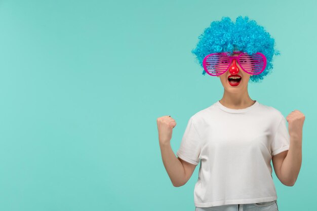 Día de los inocentes niña emocionada nariz roja despojada rosa sunglas puños feliz payaso divertido cabello azul