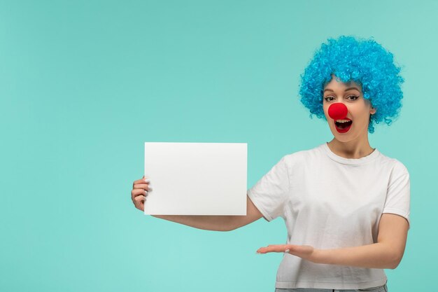 Día de los inocentes niña emocionada sosteniendo y presentando papel con nariz roja en traje de payaso cabello azul