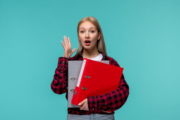 Día internacional de los estudiantes joven linda chica en camisa roja a cuadros sorprendida sosteniendo carpetas de archivos