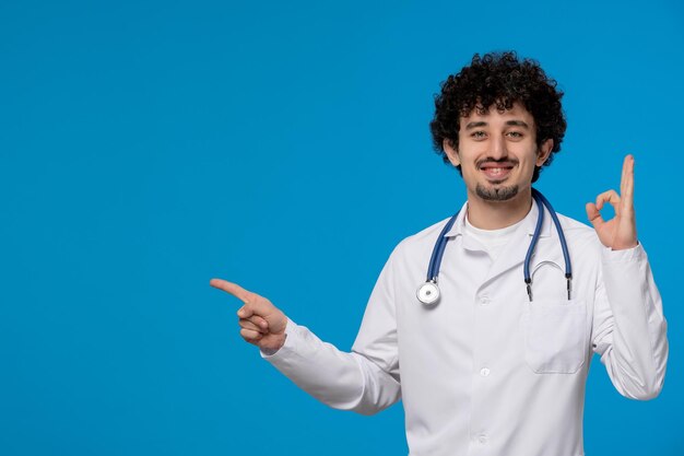 Día del médico chico lindo guapo rizado en uniforme médico que muestra gesto de signo ok