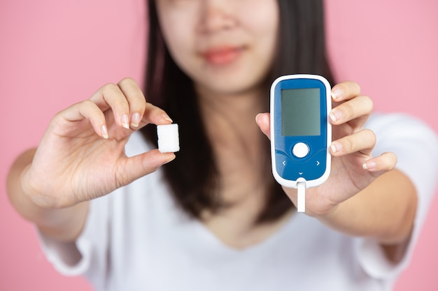 Día mundial de la diabetes; Mujer sosteniendo medidor de glucosa y terrones de azúcar en la pared rosa