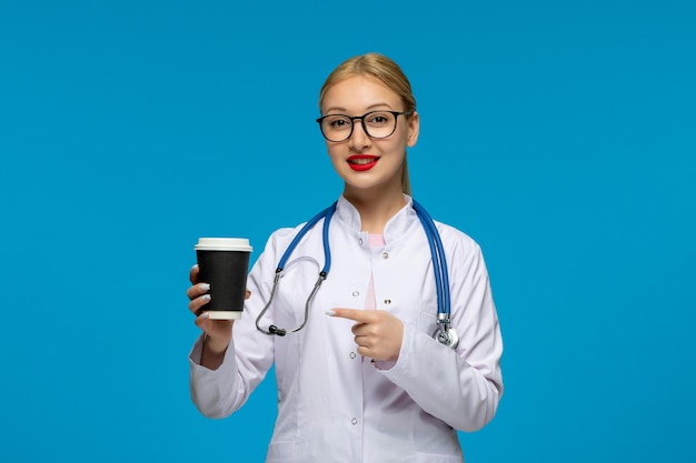 Día mundial del médico linda doctora rubia sosteniendo una taza de café con el estetoscopio en el abrigo médico