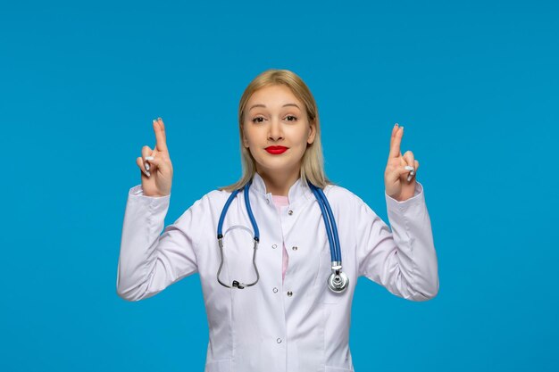 Día mundial del médico linda rubia joven médico cruzando los dedos con el estetoscopio en la bata de laboratorio