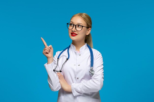 Día mundial del médico médico sonriente apuntando hacia arriba con gafas con el estetoscopio en el abrigo médico