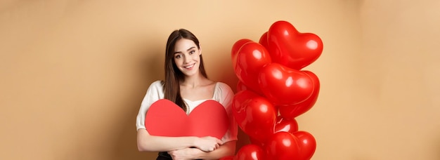 Foto gratuita el día de san valentín y el concepto de amor encantadora chica abrazando un gran corazón romántico recortado y sonriendo de pie n