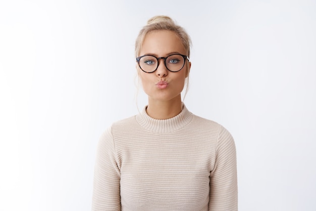 Día de San Valentín, concepto de belleza y estilo de vida. Juguetona y fllirty guapa joven europea con gafas doblando los labios enviando un beso al aire siendo romántico mostrando simpatía mwah sobre la pared blanca