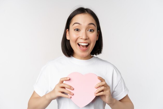 Día de San Valentín Feliz niña asiática recibe un regalo romántico sosteniendo una caja en forma de corazón y sonriendo emocionada de pie sobre fondo blanco