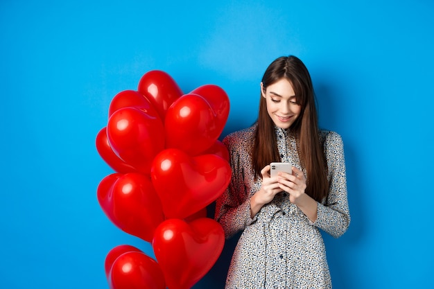 Día de san valentín. mujer sonriente en vestido de pie cerca de globos de corazones rojos y mirando smartphone, de pie sobre fondo azul.