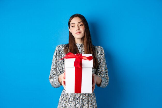 Día de San Valentín novia romántica traer regalo y sonreír a la cámara de pie en vestido de moda en azul b ...