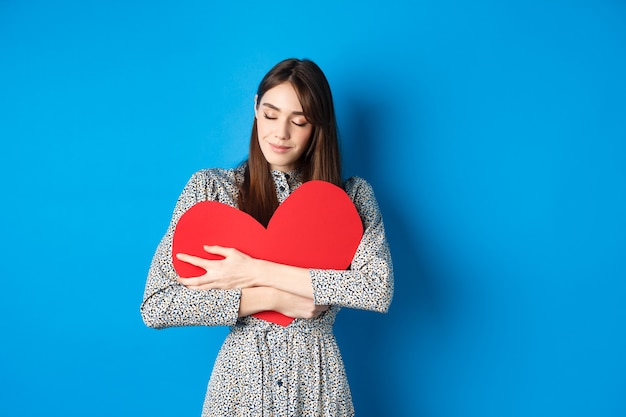 Foto gratuita día de san valentín romantinc chica en vestido abrazando gran corazón rojo recorte cerrar los ojos y sonreír con ensueño ...