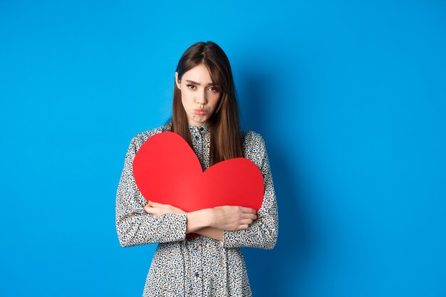 Día de san valentín triste y triste niña caucásica fruncir los labios y mirando decepcionado sosteniendo un gran corazón rojo ...