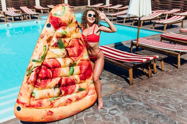 Día soleado. Bastante joven vistiendo traje de baño rojo de pie junto a la piscina, sosteniendo pizza de colchón inflable. Insta foto.