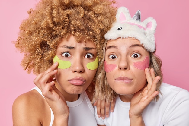 Foto gratuita día de spa mujeres jóvenes y amigables conmocionadas aplican parches de belleza debajo de los ojos para la cara táctil antienvejecimiento miran suavemente a la cámara vestidas con ropa doméstica informal aislada sobre fondo rosa