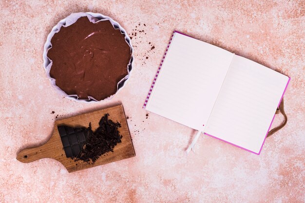 Un diario blanco en blanco abierto con la torta y la barra de chocolate quebrada en la tajadera sobre el fondo texturizado