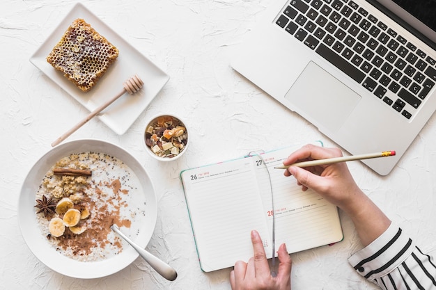 Diario de escritura de empresaria con lápiz en lugar de trabajo con ordenador portátil y desayuno saludable