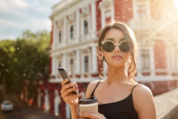 Días de verano. Ciérrese encima del retrato de la mujer caucásica femenina flaca atractiva con el pelo oscuro en anteojos bronceados y el vestido negro, esperando al novio que llega tarde a la cita, charlando con un amigo