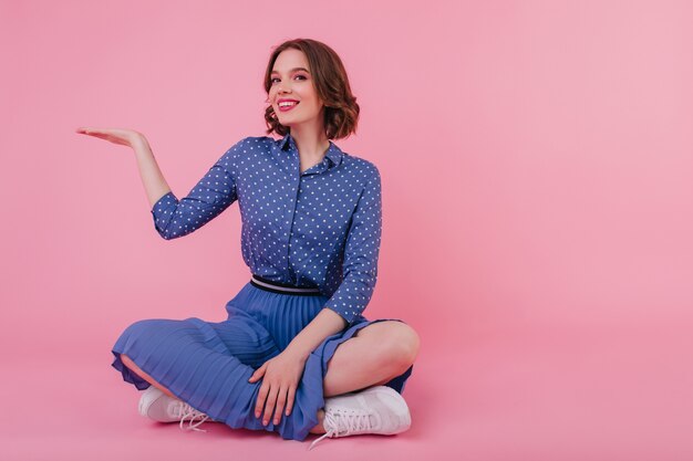 Dichosa chica de cabello castaño sentada en el suelo y sonriendo. feliz mujer morena viste blusa y falda azul.