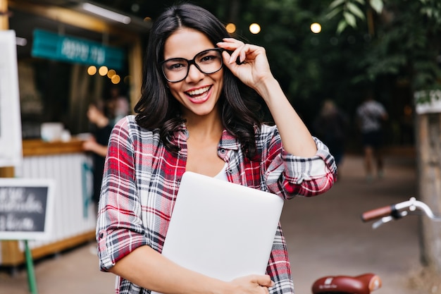 Foto gratuita dichosa chica de cabello oscuro con portátil tocando sus gafas. retrato al aire libre de freelancer latina feliz.