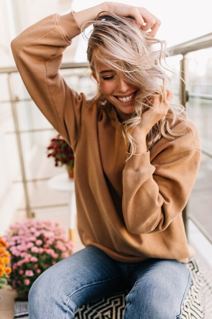 Dichosa chica en jeans riendo con los ojos cerrados. mujer joven jugando con su pelo rizado.