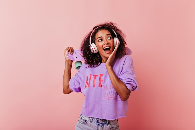 Foto gratuita dichosa chica negra con peinado rizado posando con patineta. modelo de mujer africana de buen humor en ropa casual escuchando música.